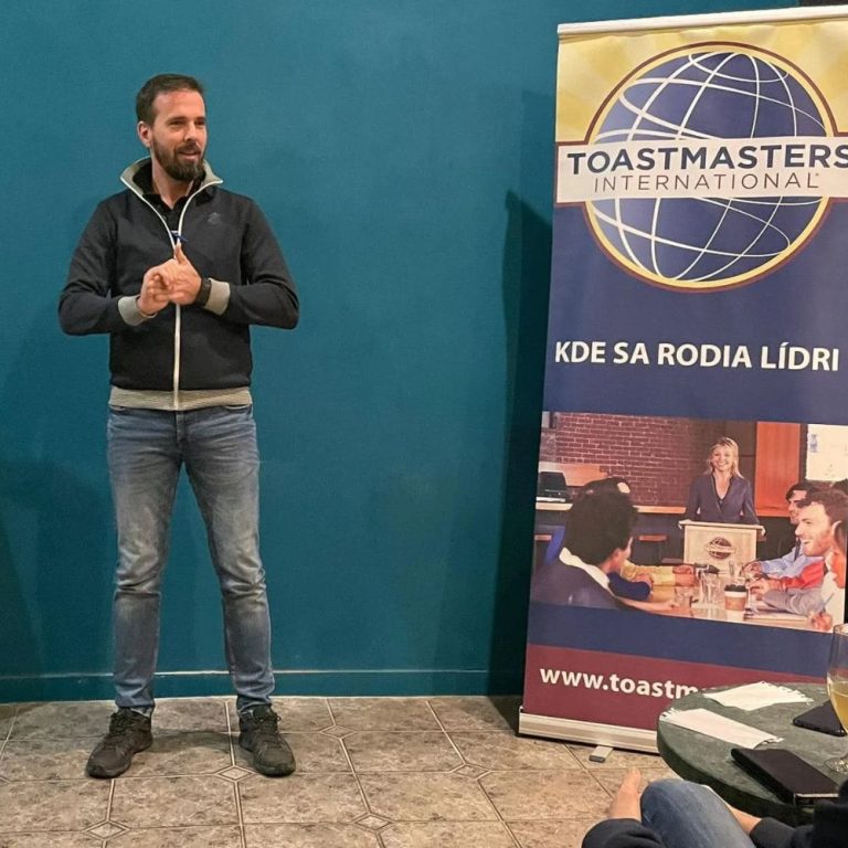 Marek Dzurilla hovorí bez prípravy pred publikom na stretnutí Toastmasters, za ním roll-up Toastmasters International s nápisom Kde sa rodia lídri