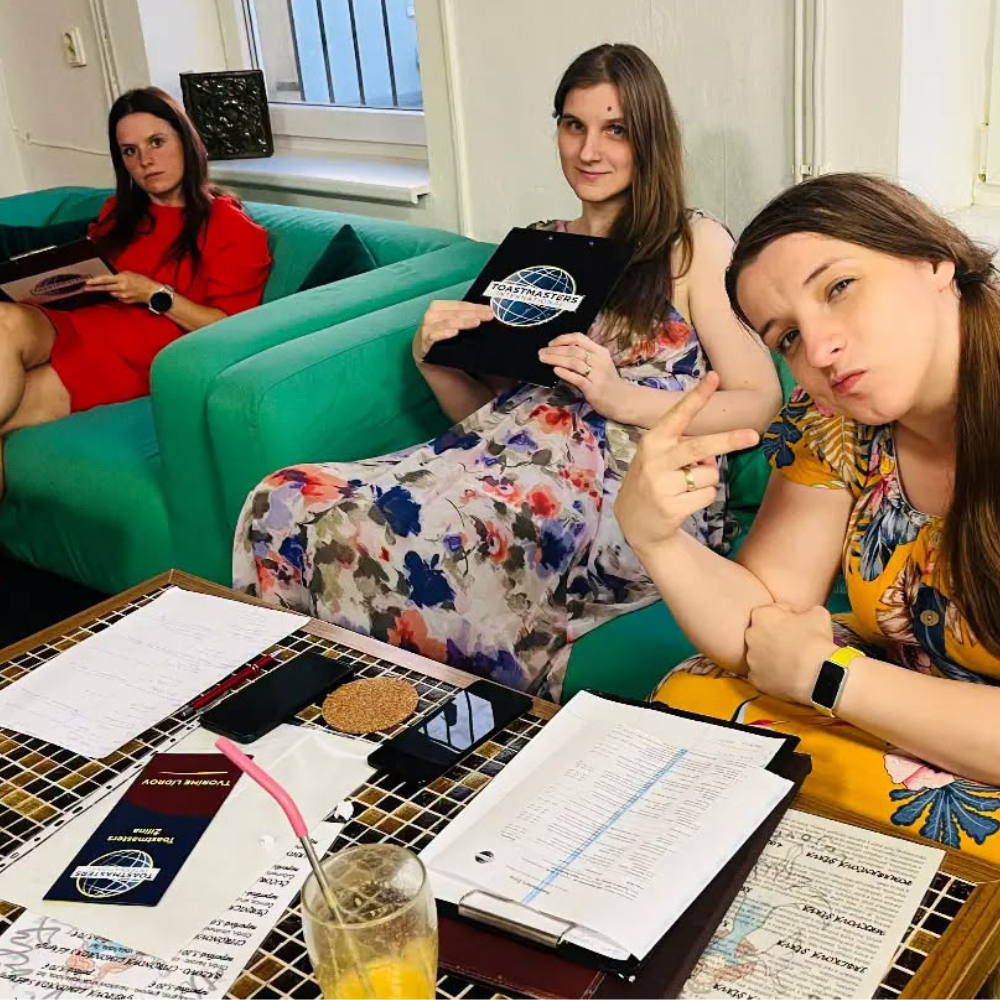 Tri členky Toastmasters na zelených sedačkách pri stole s programom stretnutia, poznámkami a deskami s logom Toastmasters, jedna z nich žartovne pózuje do objektívu